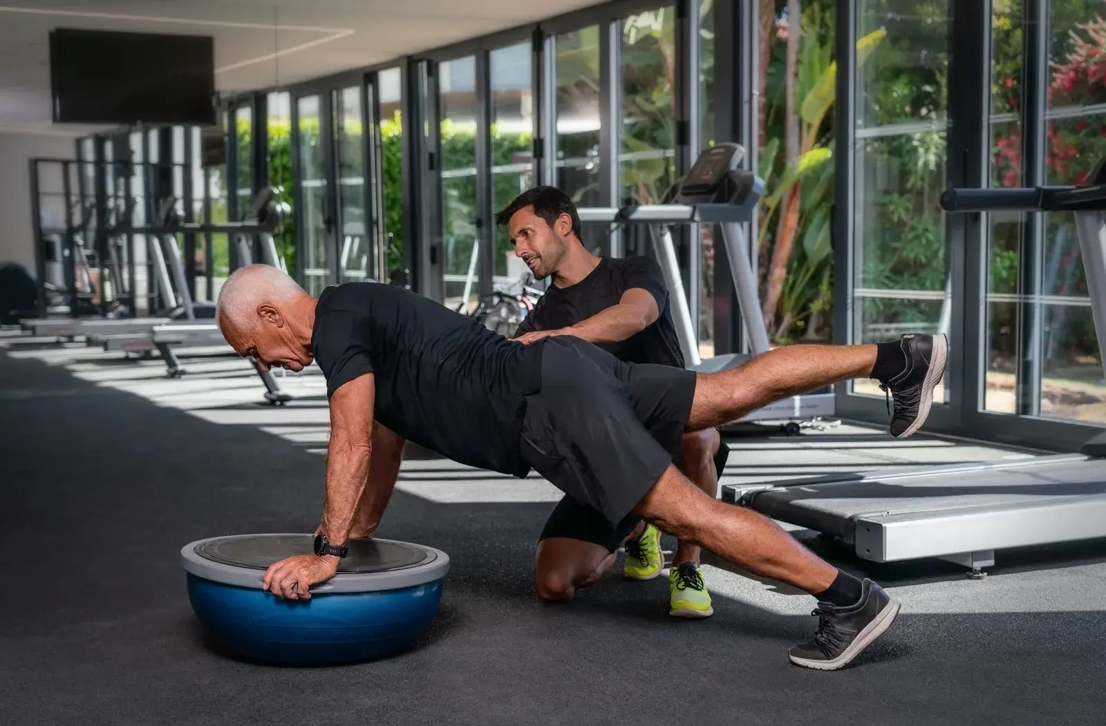 A man holds a plank on a balance trainer while a coach kneels beside him