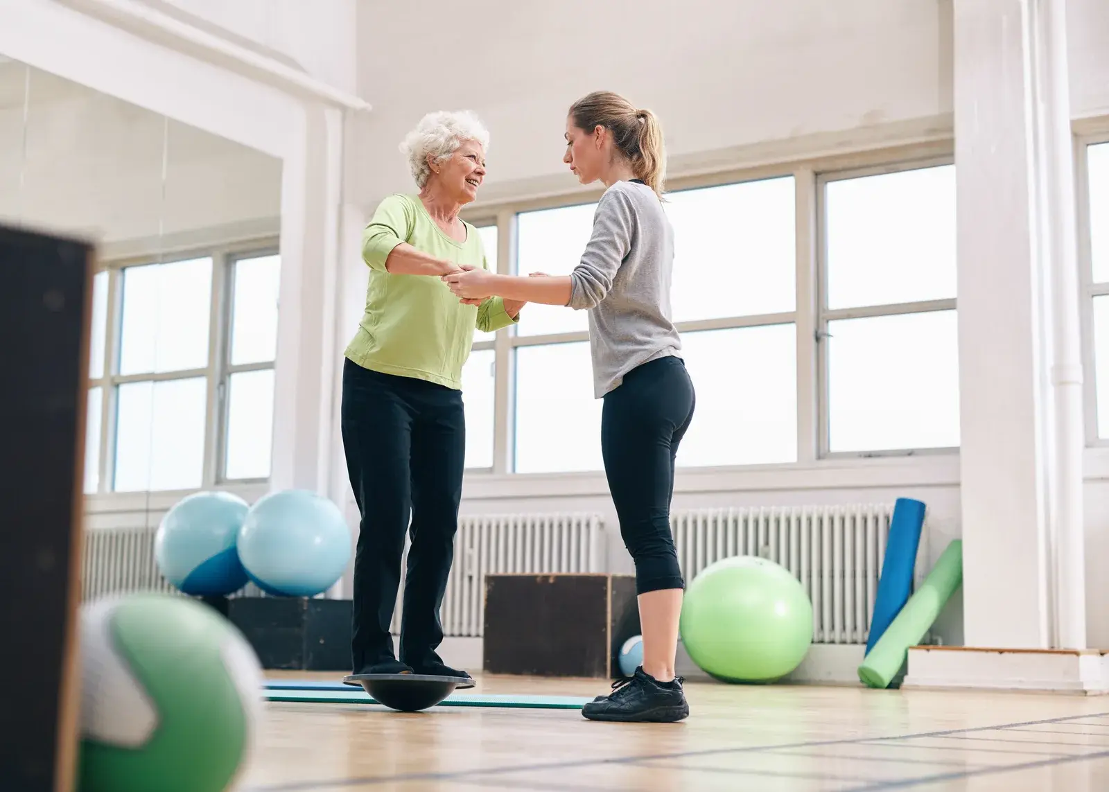An older woman practices balance while holding an instructor’s hands in a bright studio