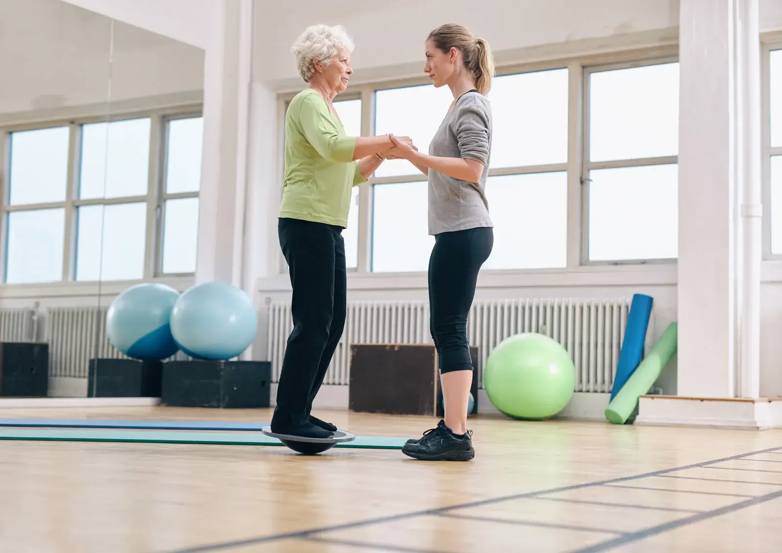 An older woman standing on one leg on a mat with an instructor supporting her.