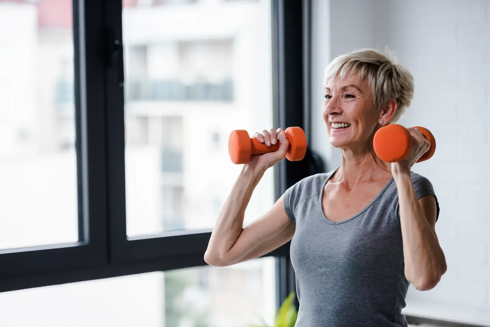 A smiling older woman lifting light dumbbells overhead beside a window.