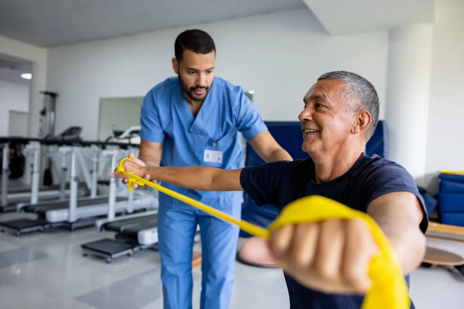 A clinician in blue scrubs guides a middle-aged man pulling a yellow resistance band
