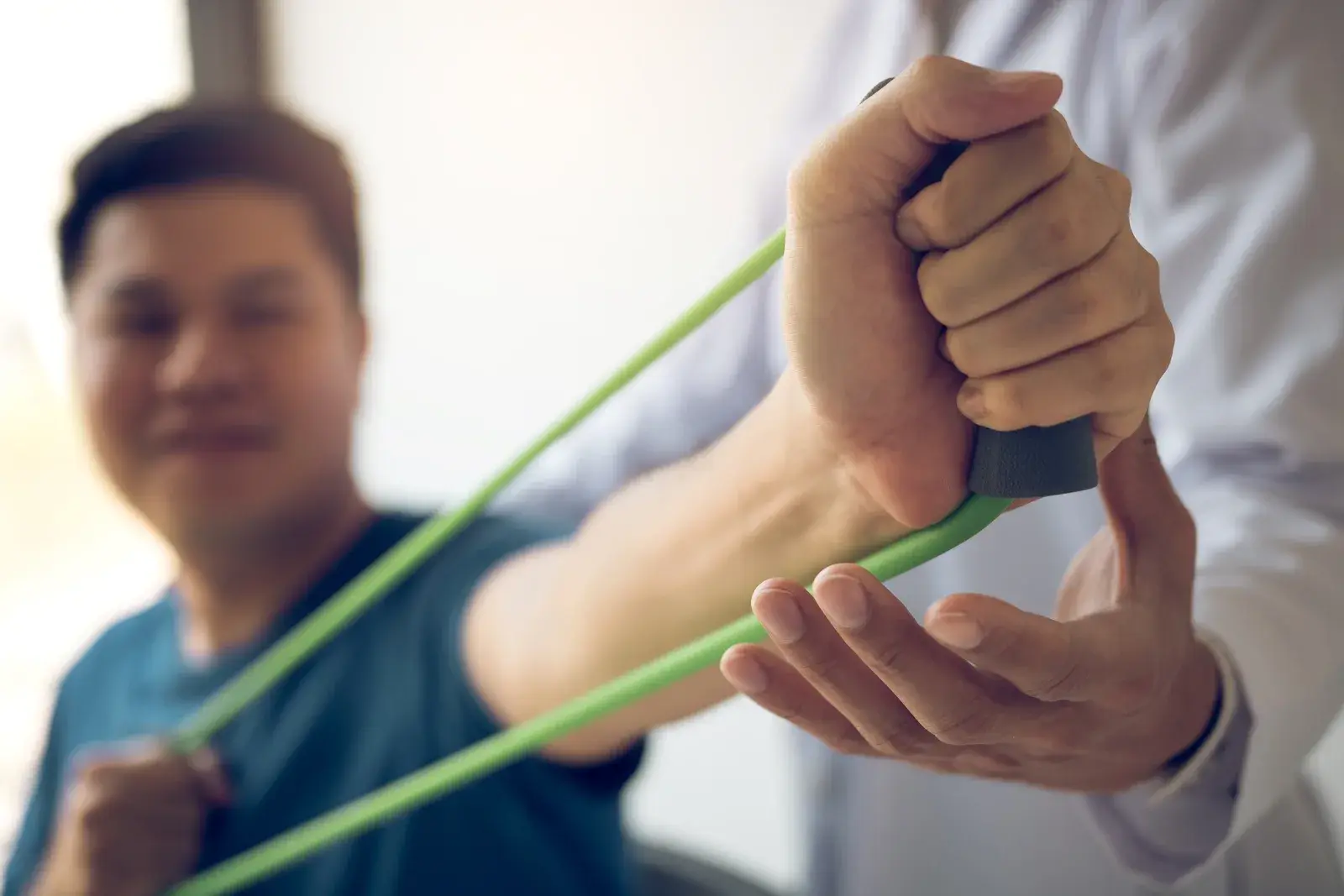 A man pulls a green resistance band while another person’s hand steadies his forearm