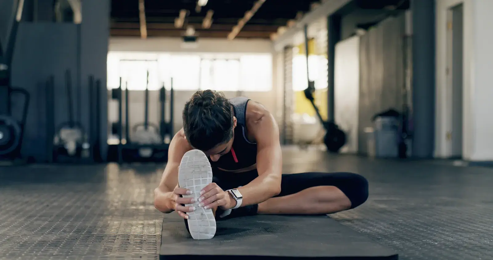 A person seated on a gym floor reaching toward one foot in a hamstring stretch
