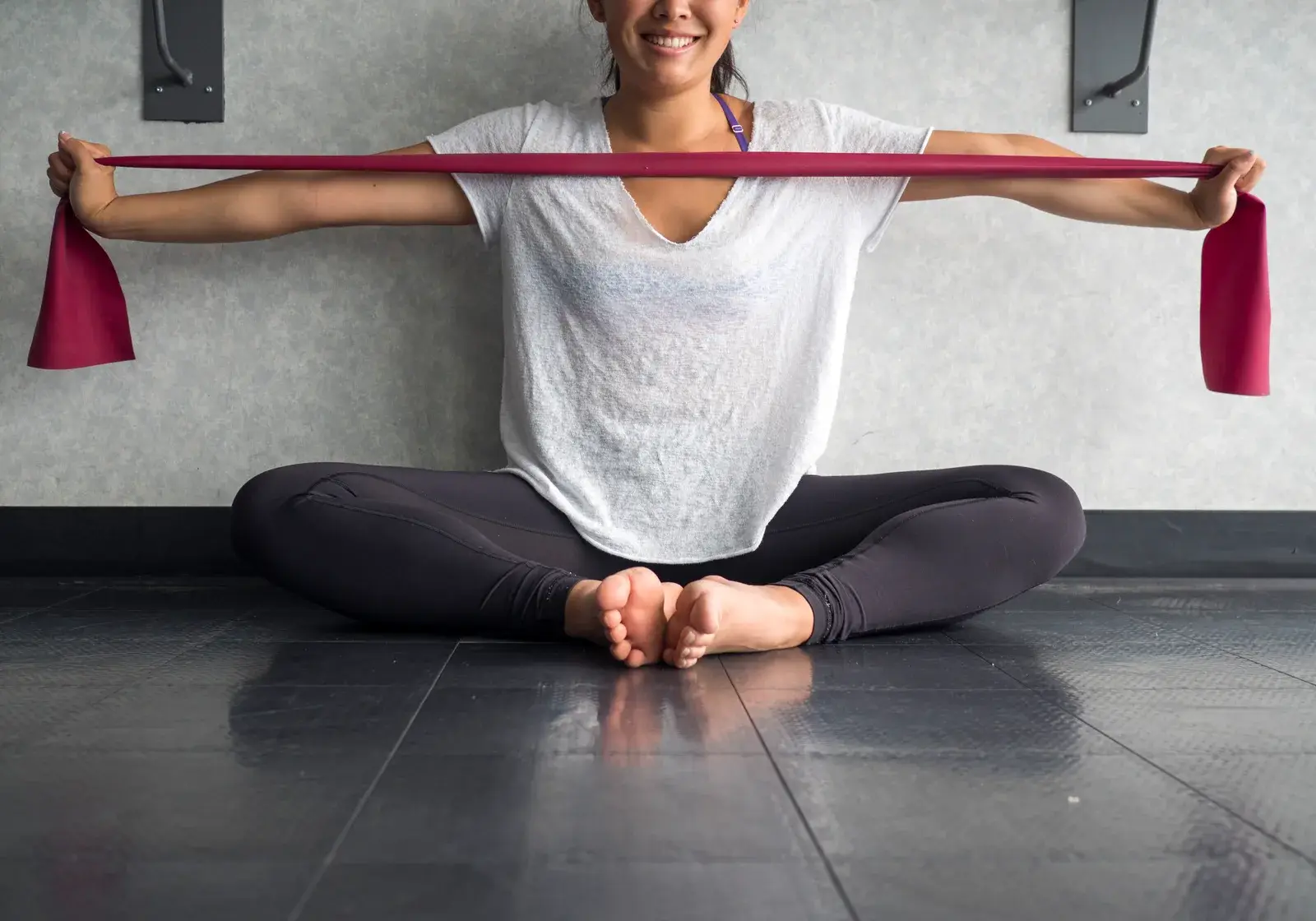 A woman seated on a studio floor pulls a resistance band apart at chest height