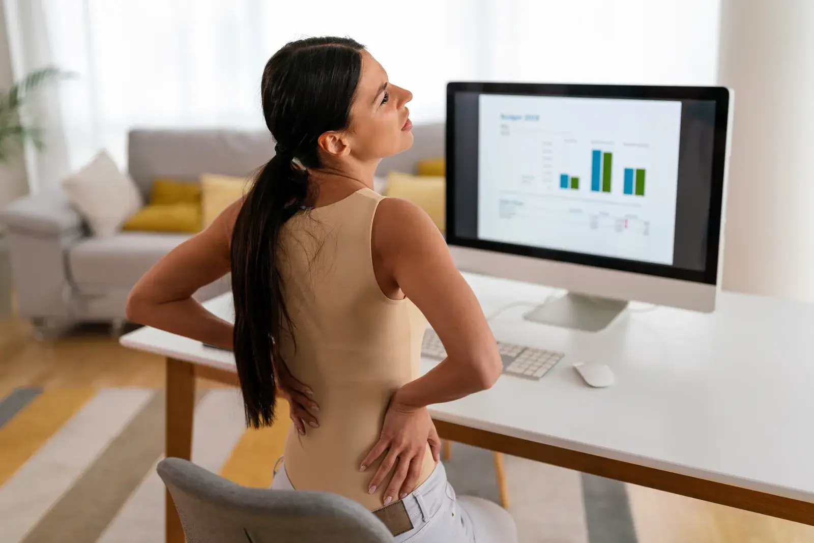 A woman seated at a computer desk presses both hands into her lower back